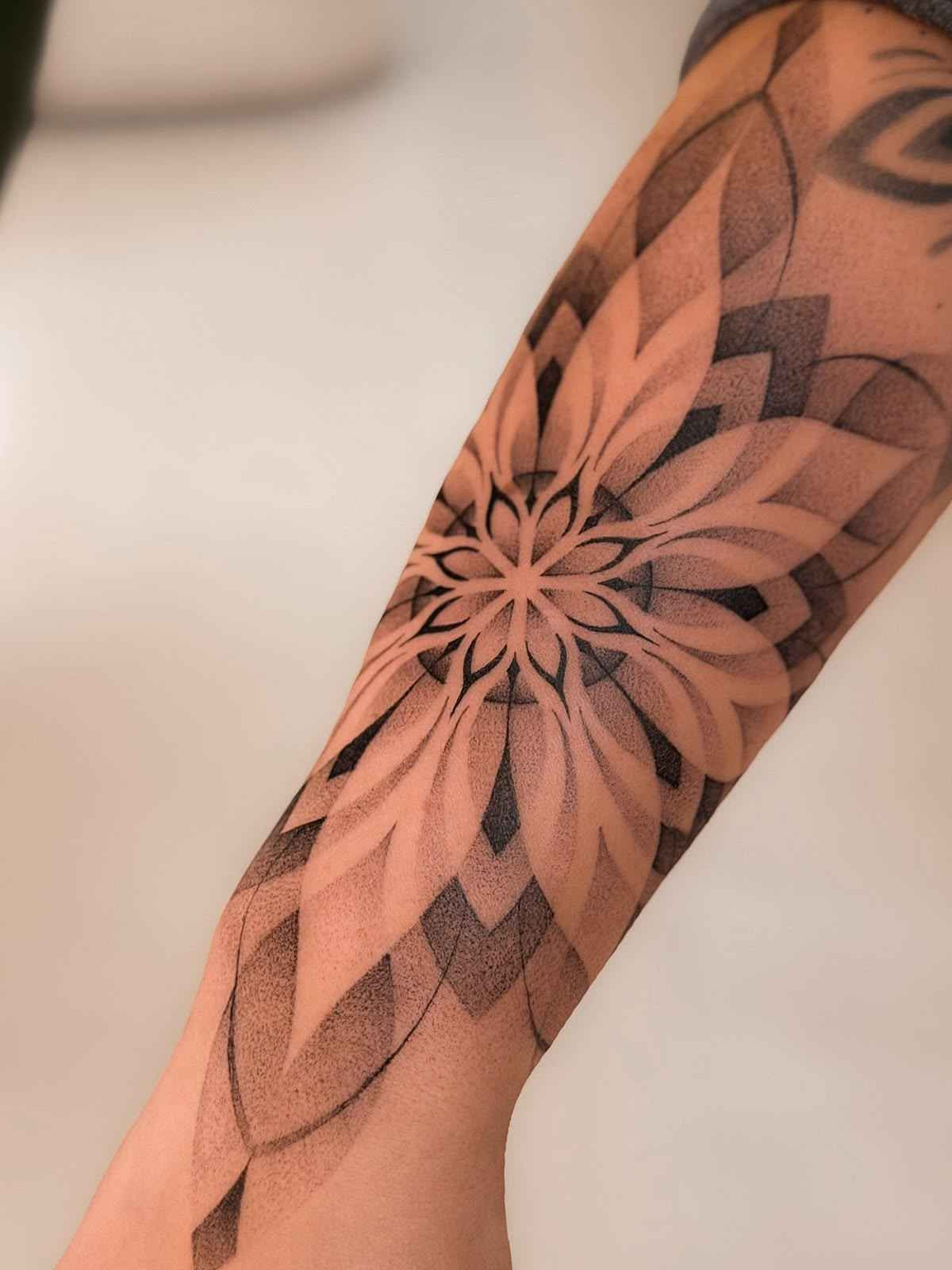 Ornamental tattoo featuring a layered mandala flower with geometric patterns and soft dotwork shading on the forearm