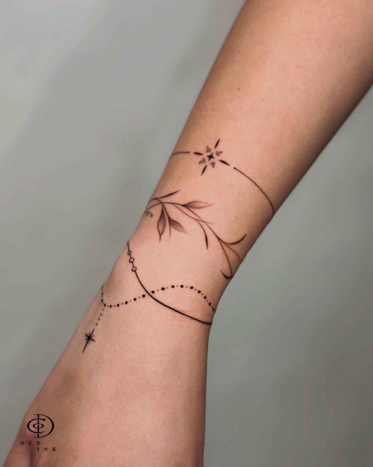 ornamental tattoo featuring delicate bracelet lines, botanical elements, and ornamental dots wrapped around the forearm