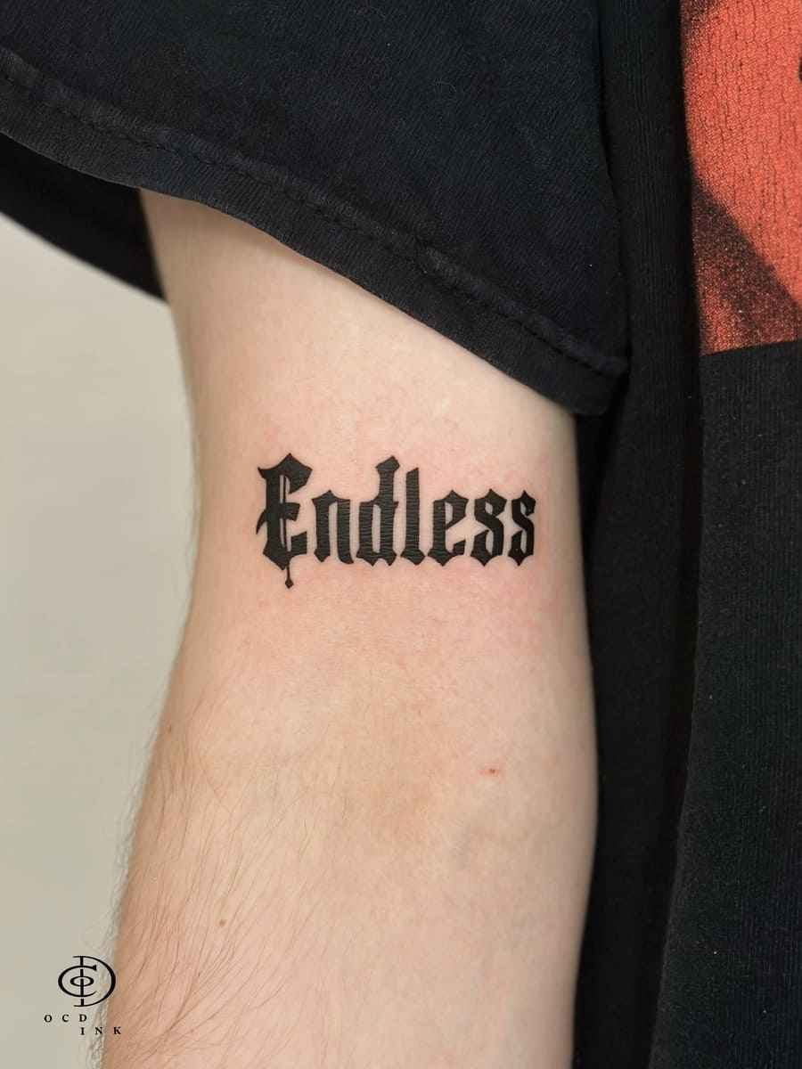 Gothic lettering tattoo featuring bold Old English typography in solid black ink on the arm