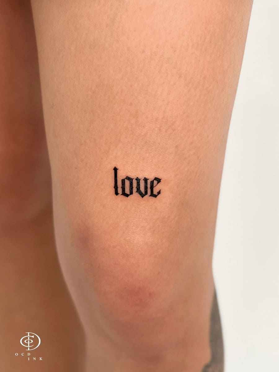 Small Gothic lettering tattoo reading "love" in Old English font on the inner arm