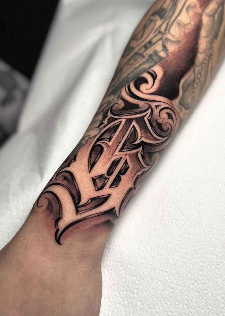 Chicano lettering tattoo featuring an ornate Gothic-style initial with black and gray shading on the forearm