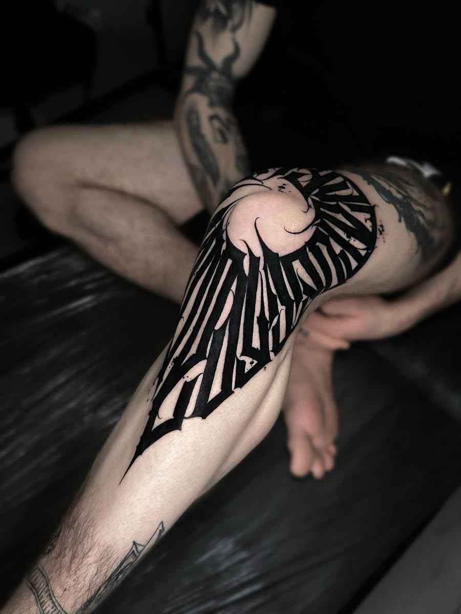 Large Gothic lettering tattoo with bold black Old English typography wrapping around the knee