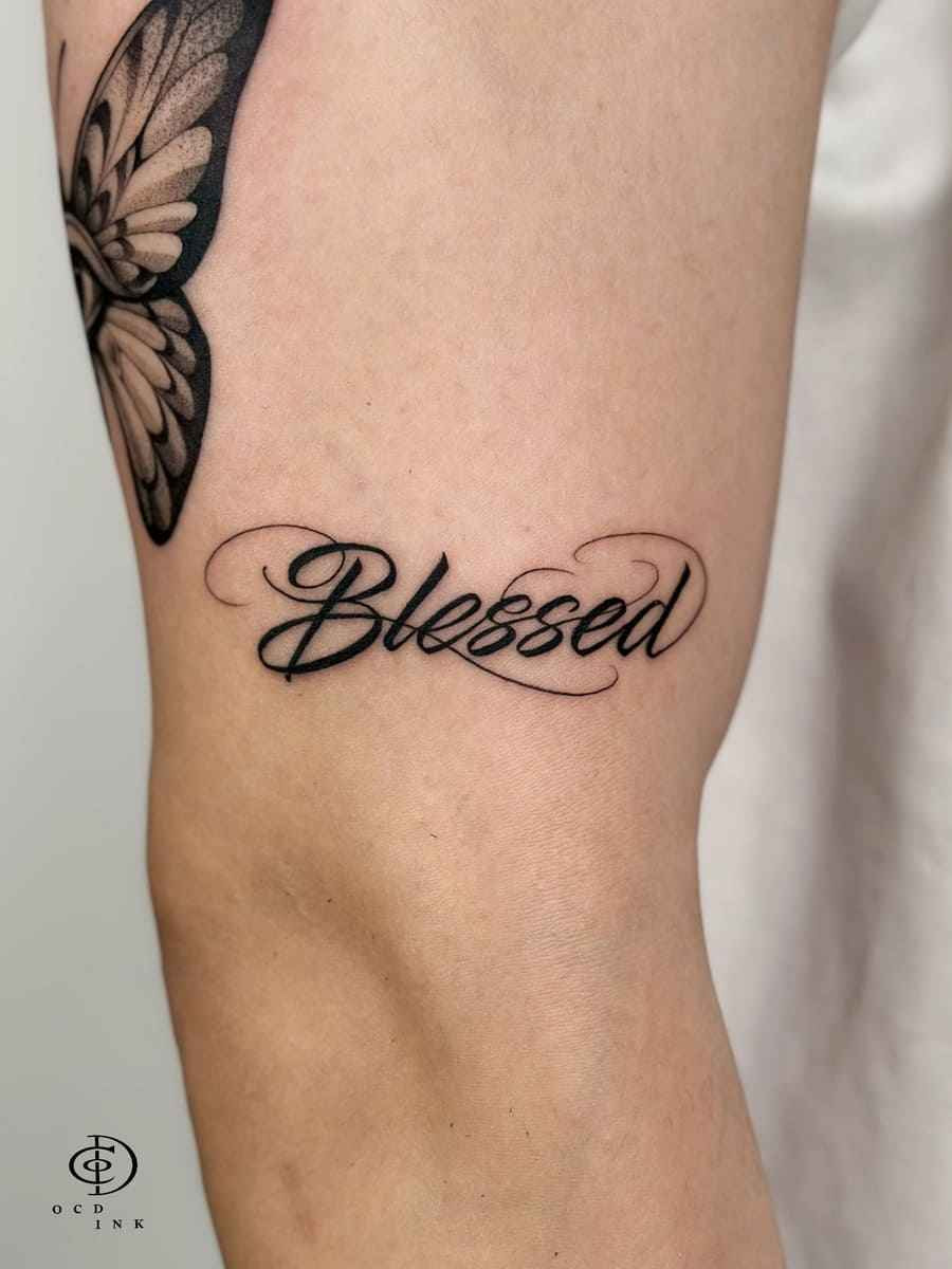 Script calligraphy tattoo reading "Blessed" with elegant cursive lettering on the leg