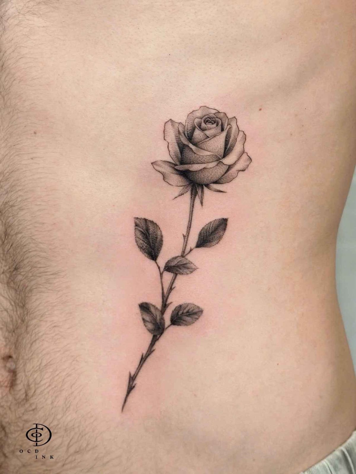 Black and grey dotwork tattoo of a single blooming rose on the ribcage, featuring realistic floral details, fine linework, and smooth stipple shading.