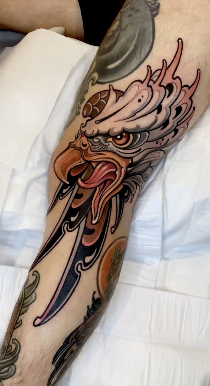 COLOR Neo traditional color tattoo of an eagle head wrapped around a dagger, featuring bold outlines, expressive details, and warm brown, cream, and red tones.