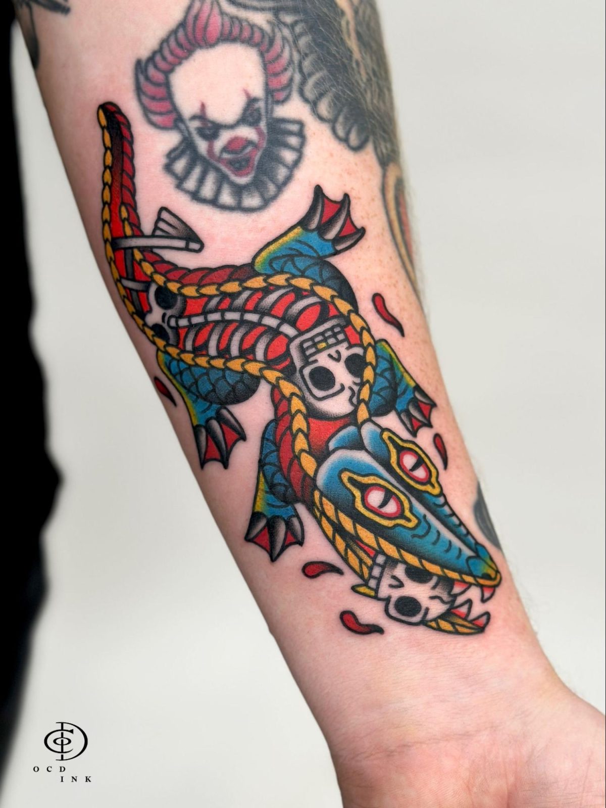 COLOR American traditional tattoo of a blue crocodile wrapped in rope with skulls, featuring bold black outlines, saturated red, blue, yellow, and green colors.