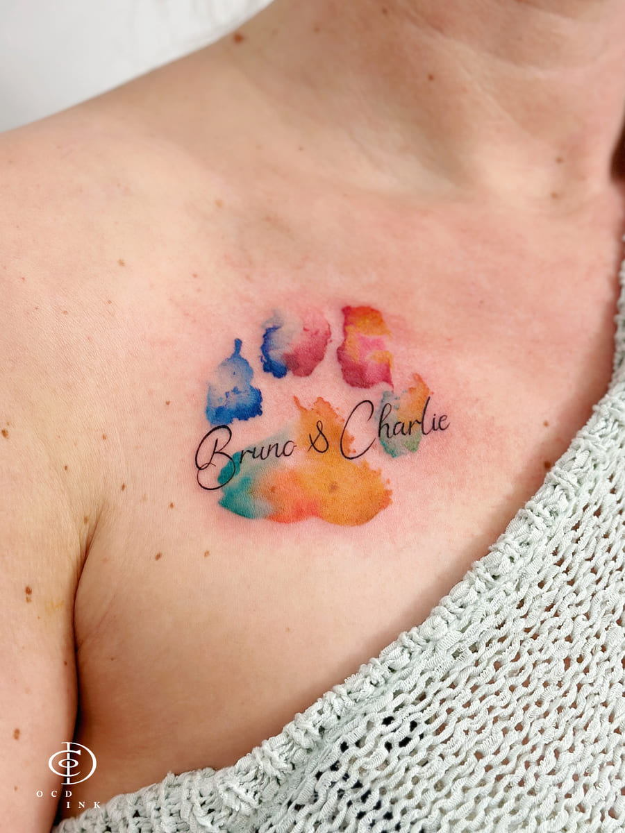 COLOR Watercolor tattoo featuring colorful dog paw prints with the names Bruno and Charlie in elegant script, blending soft blue, pink, orange, and green watercolor effects.