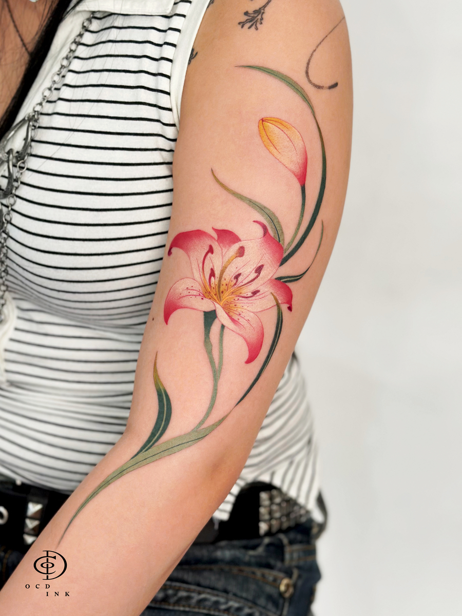 COLOR color tattoo featuring a blooming pink lily with a yellow flower bud and flowing green leaves, elegantly placed along the upper arm.