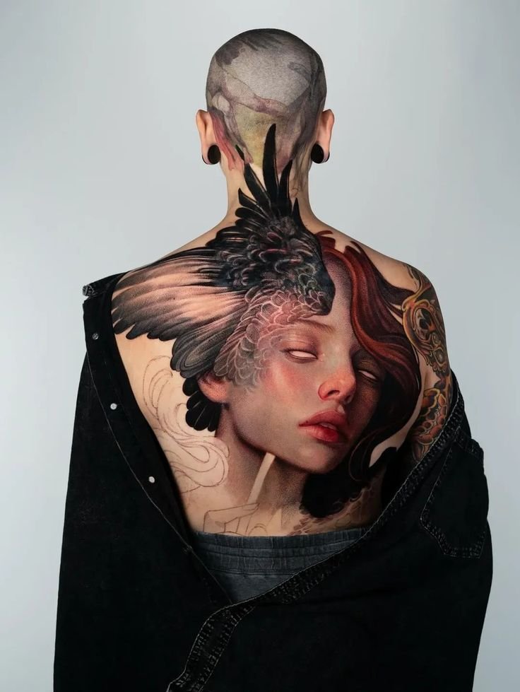 Large surrealism tattoo covering the upper back, featuring a realistic female portrait seamlessly blended with black bird wings and abstract dreamlike elements in a full-color composition.
