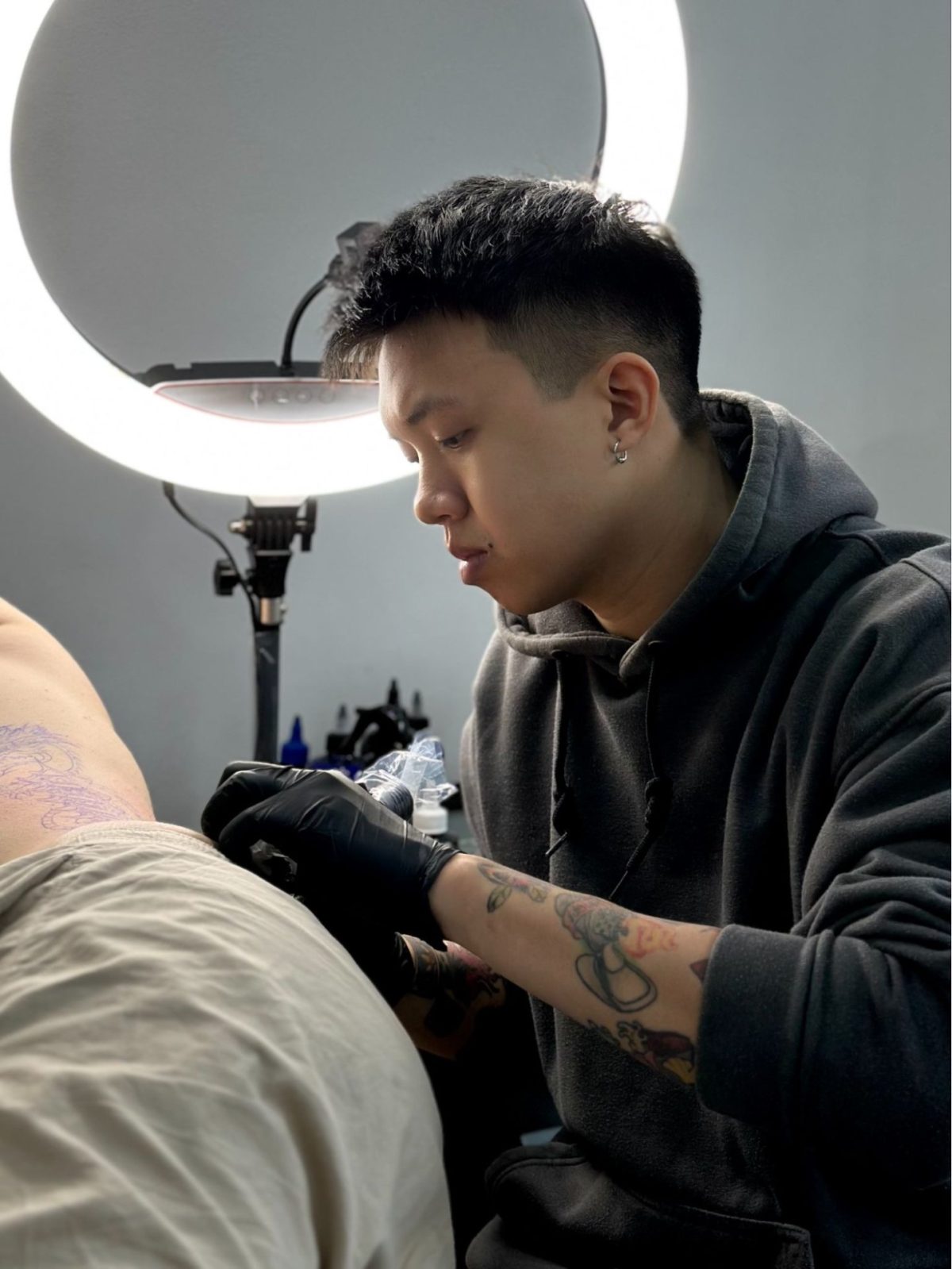 A tattoo artist wearing a dark hoodie and black nitrile gloves carefully tattoos a client under a bright circular ring light