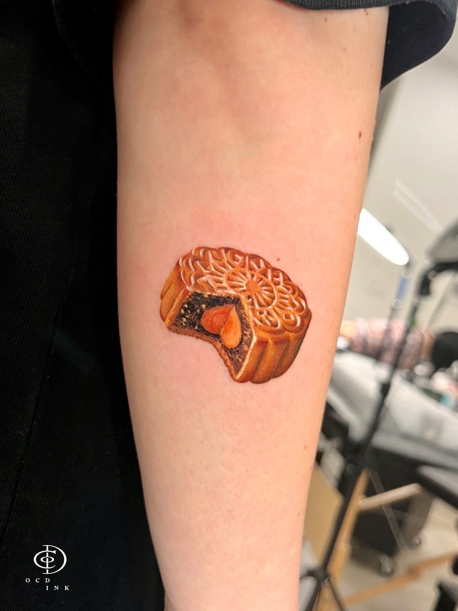 Micro realism tattoo of a traditional mooncake with a sliced section revealing a golden salted egg yolk filling, rendered with realistic textures and warm golden-brown tones.