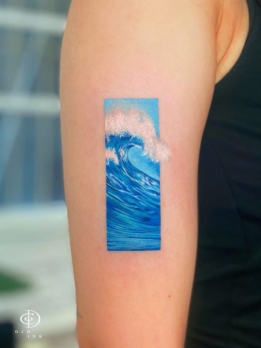 Fine art color tattoo of a crashing ocean wave inside a vertical rectangular frame, featuring vivid blue tones, detailed white sea foam, and a minimalist composition on the upper arm.