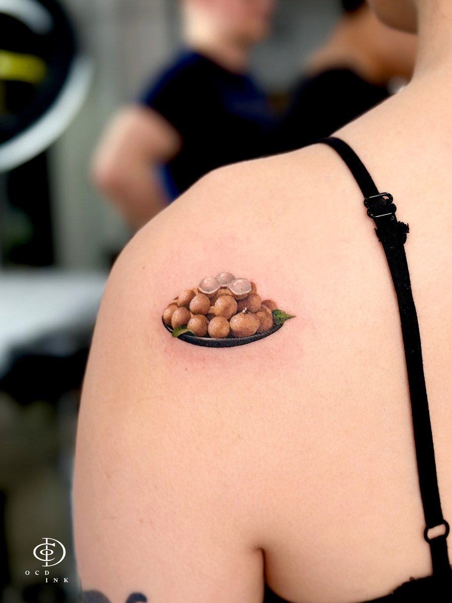Micro realism tattoo of a plate of fresh longan fruit, including peeled and whole longans with green leaves, rendered in lifelike detail on the shoulder.