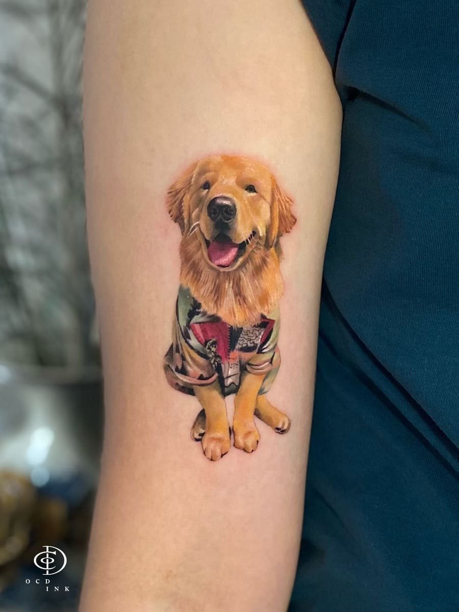 Hyper realism color tattoo of a smiling Golden Retriever wearing a patterned shirt, showcasing lifelike fur texture, expressive eyes, and realistic shading on the upper arm.