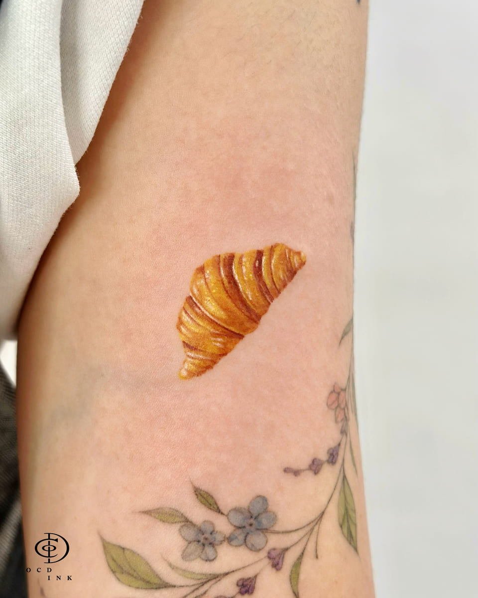 Tiny micro realism tattoo of a golden-brown croissant on the inner arm, featuring realistic flaky layers, buttery texture, and lifelike shading in a minimalist design.