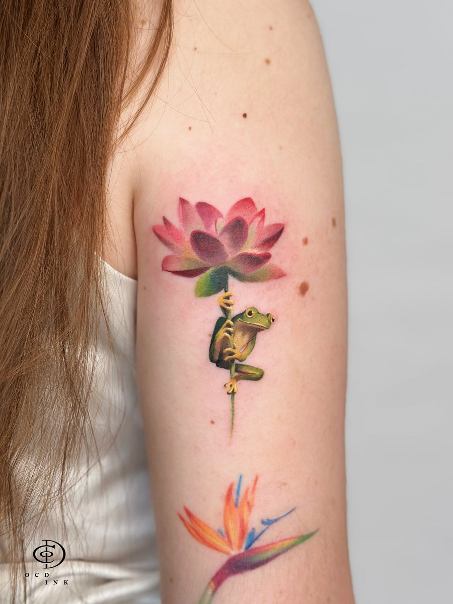 Realistic color tattoo featuring a frog climbing a pink lotus flower on the upper arm, complemented by a colorful bird-of-paradise flower below.