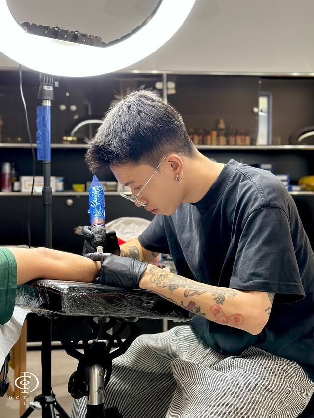 Black & Gray A professional tattoo artist wearing glasses and black gloves carefully tattoos a client's forearm in a modern studio.