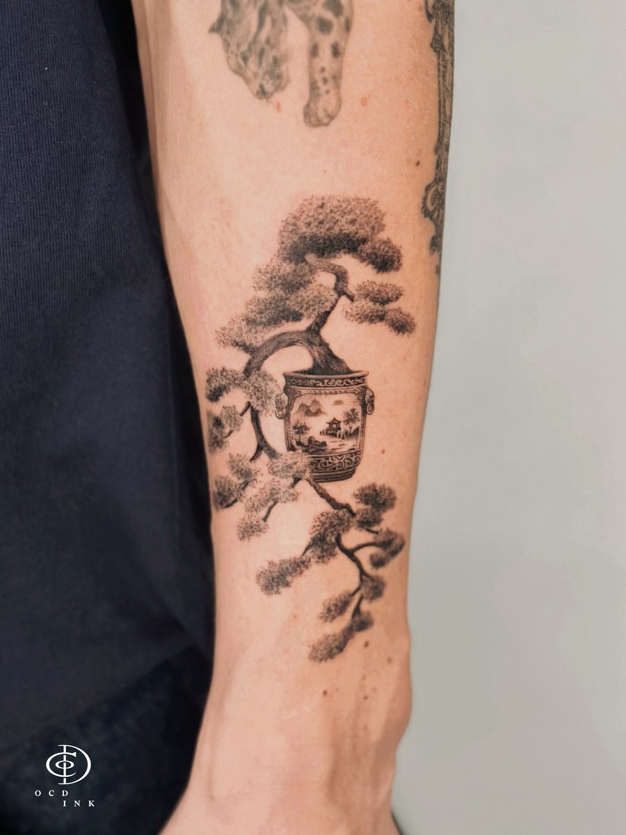 Black & Gray A black and gray bonsai tattoo featuring a delicately textured tree growing from an ornate ceramic pot. Soft stippling and fine shading give the miniature tree a traditional ink painting aesthetic on the forearm.