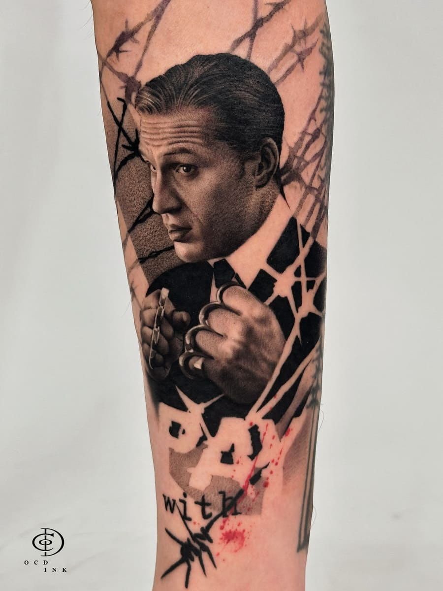 Black & Gray A black and gray realism portrait tattoo of a well-dressed man gripping brass knuckles, featuring dramatic shading, detailed facial features, and a cracked background with subtle red splatter accents for a cinematic effect