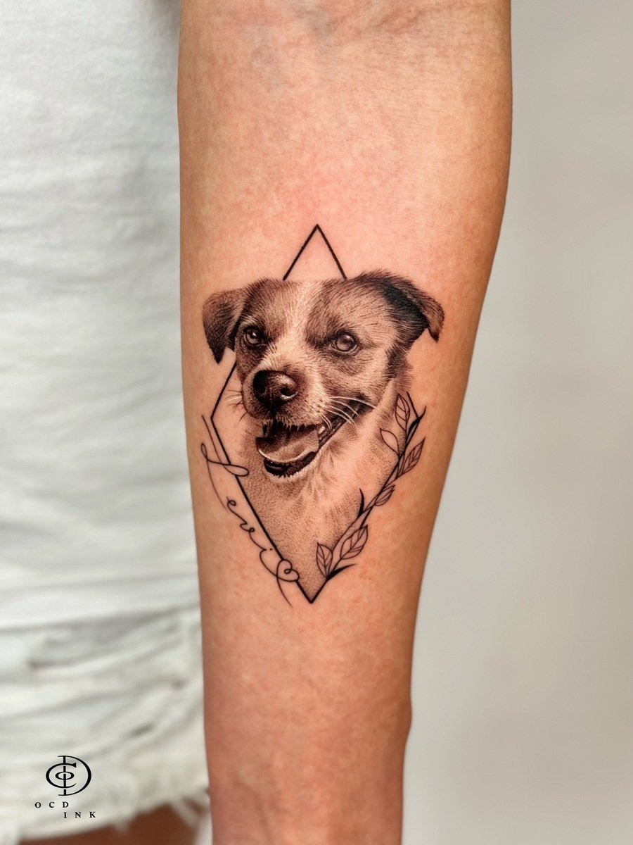 Black & Gray A black and gray pet portrait tattoo of a smiling dog framed by fine-line geometric shapes and botanical elements on the forearm