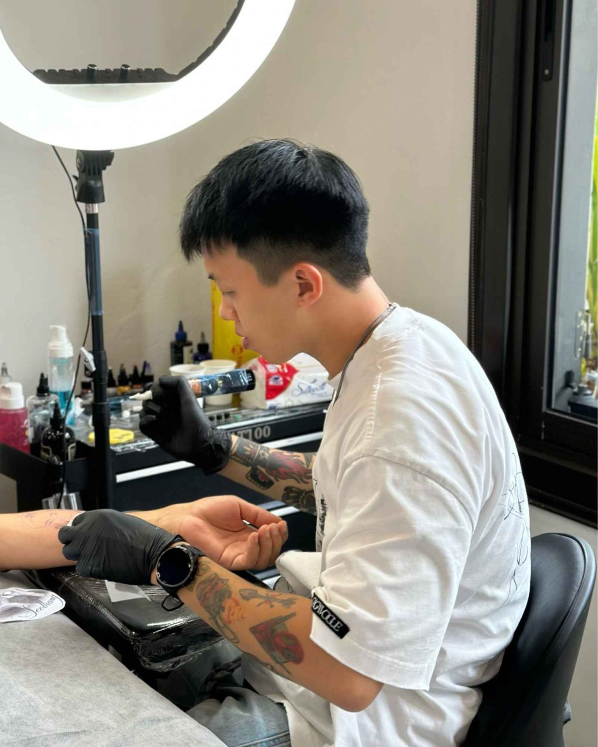 Traditional tattoo Tattoo artist wearing black gloves works carefully on a client’s forearm in a professional tattoo studio under a bright ring light.