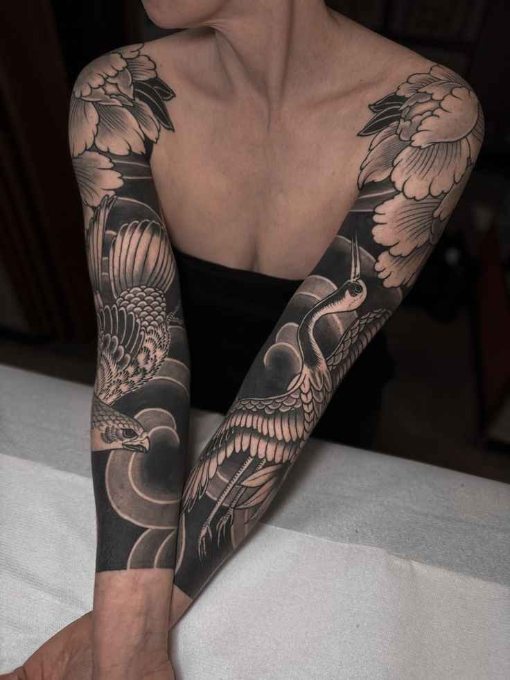 Traditional Japanese black-and-gray sleeve tattoos featuring cranes, floral motifs, and sweeping dark background patterns across both arms