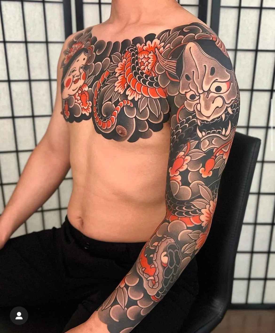 Traditional Japanese tattoo covering the chest and arm with a Hannya-style mask, serpent, flowers, and dark wave-like background patterns.