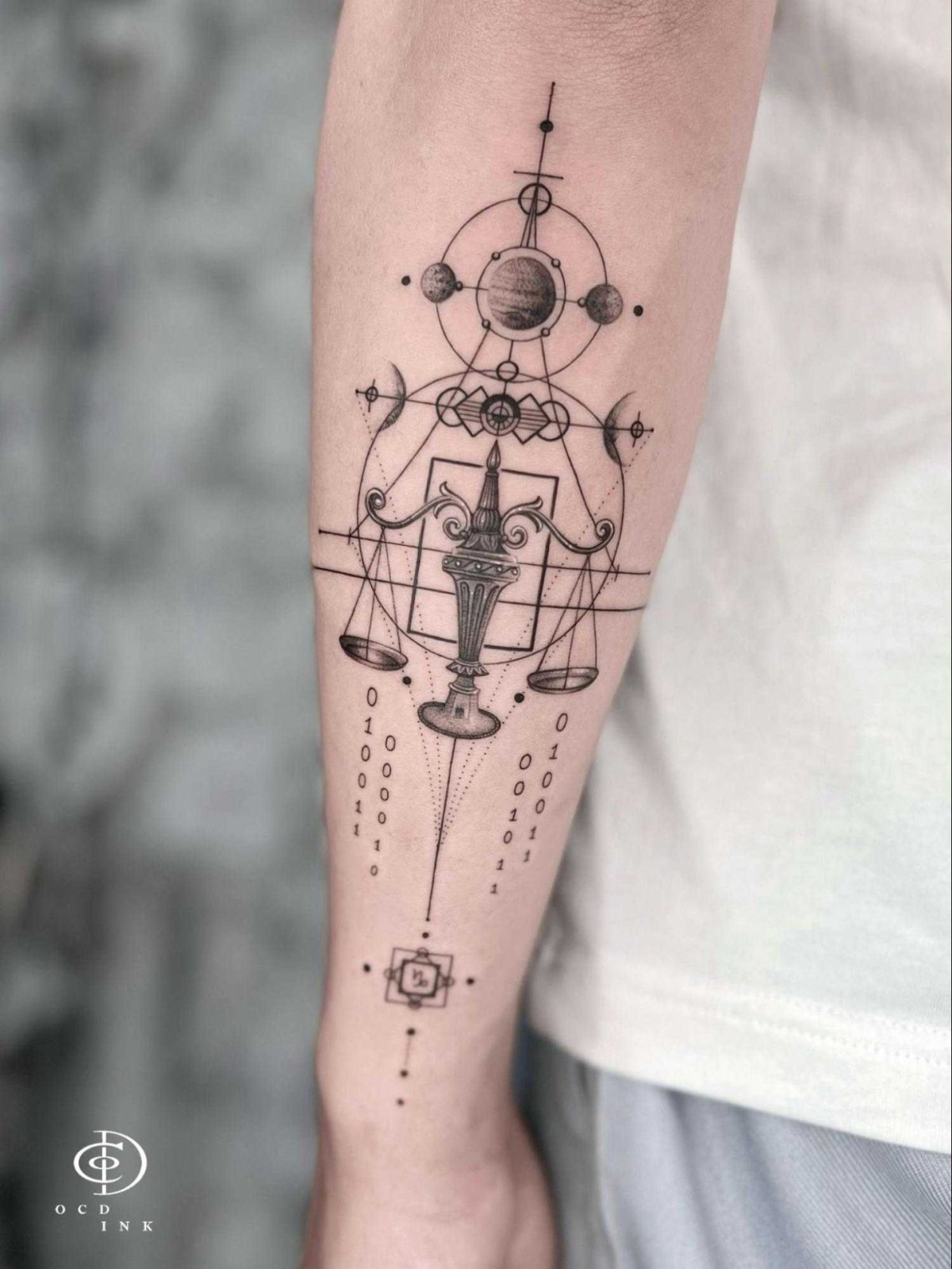 Geometric graphic tattoo with scales, orbiting planets, abstract symbols, and binary code arranged along the forearm