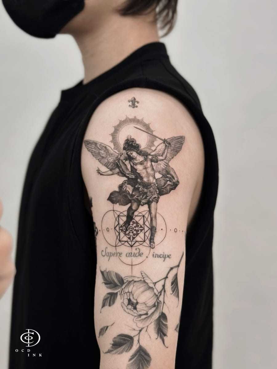 Graphic upper-arm tattoo featuring a winged warrior, geometric linework, script lettering, and a detailed botanical flower