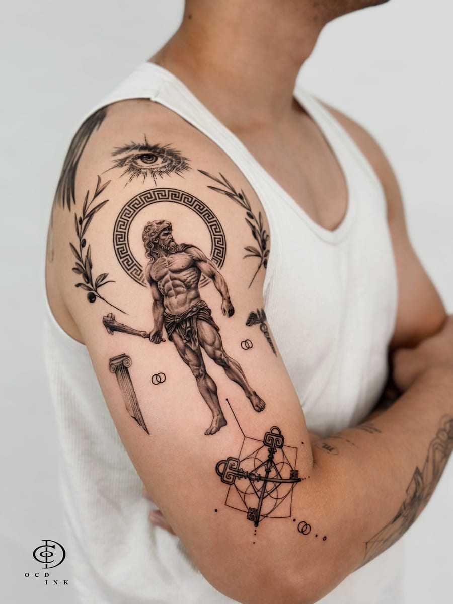 Graphic tattoo on the upper arm featuring a classical muscular figure surrounded by an eye, laurel branches, architectural fragments, and geometric symbols