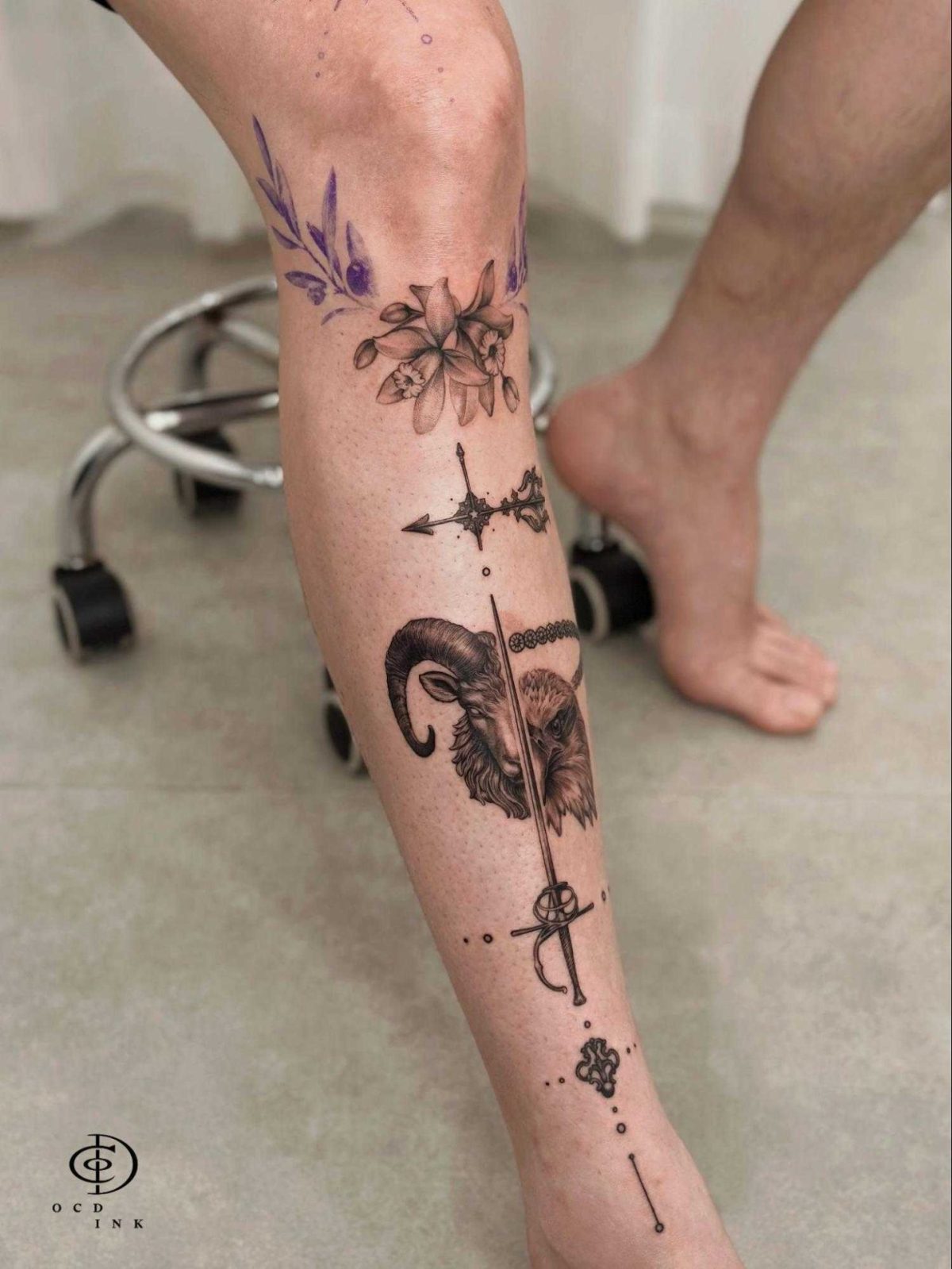 Graphic leg tattoo combining a ram and owl portrait with a vertical sword, ornamental details, and delicate floral elements