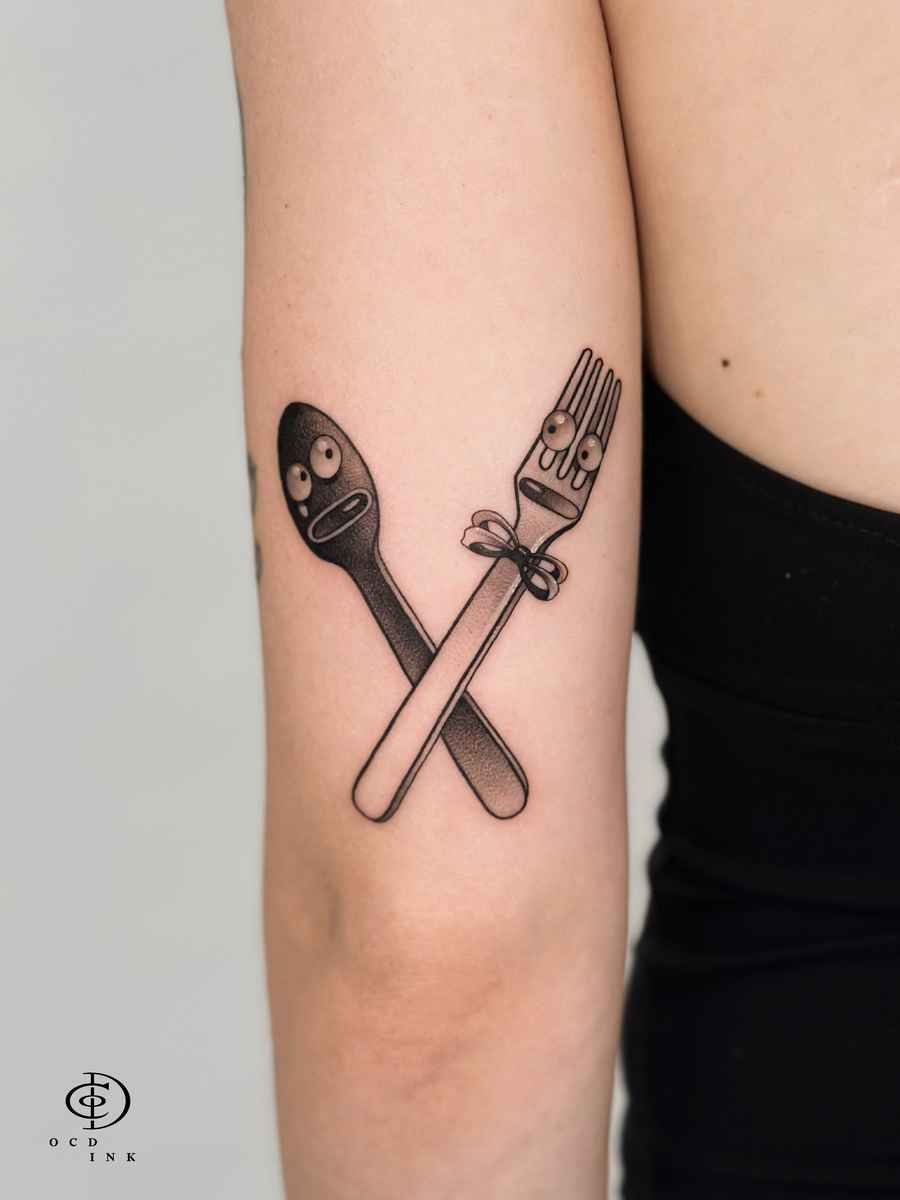 ILLUSTRATION Illustration tattoo of a crossed spoon and fork with expressive eyes and faces on the upper arm