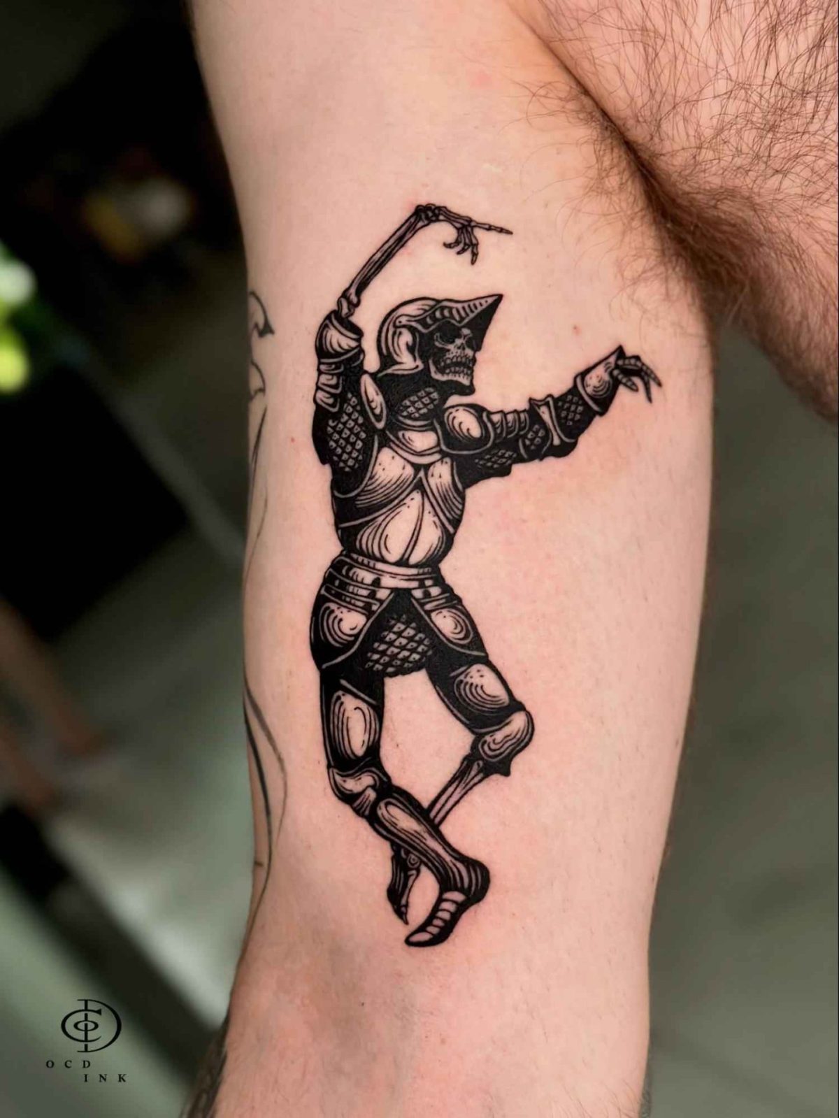ILLUSTRATION Illustration tattoo of a skeletal knight in detailed medieval armor posed dramatically on the upper arm.