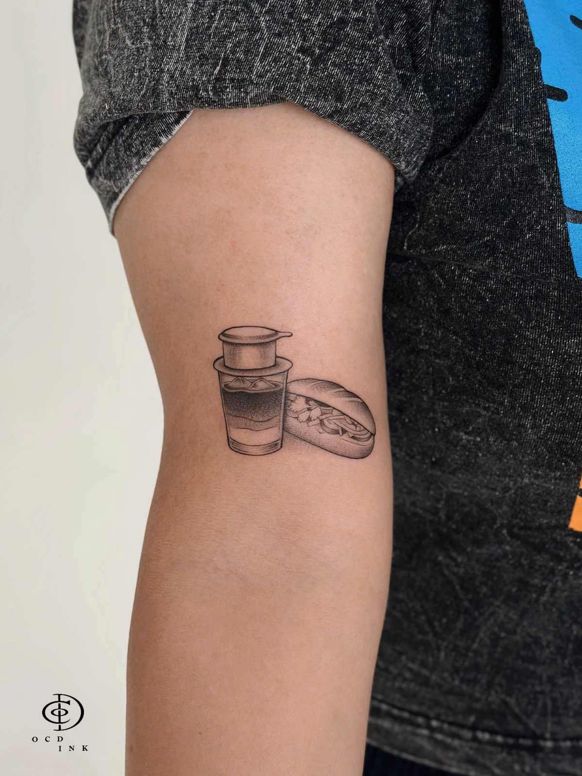 Black and grey Vietnam souvenir tattoo featuring a Vietnamese coffee cup and banh mi sandwich, two familiar symbols of Vietnamese street food culture. The tattoo combines realistic details, soft black and grey shading, and carefully placed linework to capture the charm of a simple Vietnamese breakfast tattoo on the upper arm by OCD Ink Studio in Hoi An
